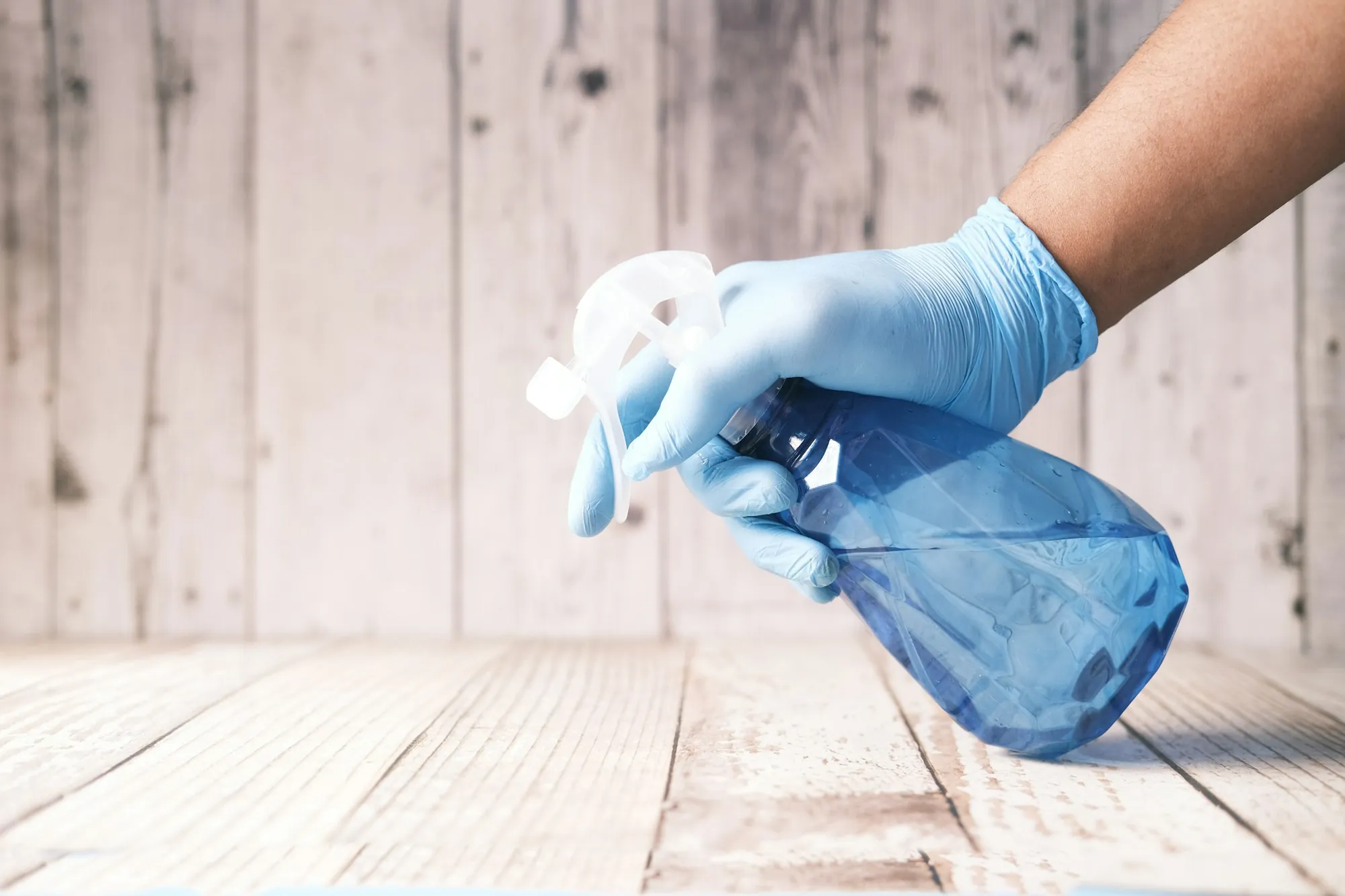 Gloved hand holding a blue spray bottle against a pale wood wall